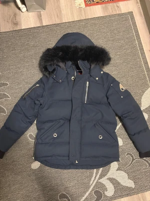 Mörkblå dunjacka Moose Knuckles - Säljer en mörkblå dunjacka från Moose Knuckles med svart päls på huvan, coola metalldetaljer och dragkedjor. Jackan har broderad hockeypatch på insidan och flera fickor med dragkedja. Perfekt för kalla dagar och riktigt snygg stil.