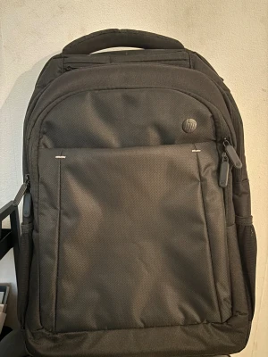 Svart ryggsäck från HP - Snygg svart ryggsäck från HP med flera fack och vadderade axelremmar. Perfekt för att bära laptop och skolgrejer. Tillverkad i slitstark polyester med dragkedjor och HP-logga framtill. Ryggpanelen är ventilerad för extra komfort.