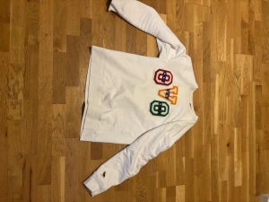 Vit OVO sweatshirt med färgglada patches - Lägg bud Säljer en vit sweatshirt från OVO med broderade patches i rött, gult och grönt framtill. Tröjan har lång ärm och klassisk passform. Tillverkad i mjuk bomull och har OVO:s ikoniska ugglelogga på ärmen. Perfekt för dig som gillar streetwear med färg.