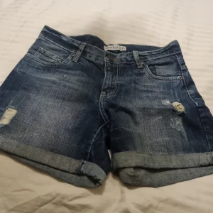 Blå denimshorts från Denim36 - Snygga blå jeansshorts från Denim36 med klassisk femficksdesign, slitna detaljer och uppvikta benslut. Shortsen har normal passform och är gjorda i bomull för en skön känsla. Perfekta för sommarens alla äventyr!