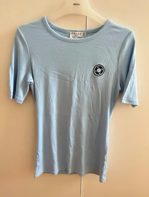 Ljusblå t-shirt från FRAME - Ljusblå t-shirt från FRAME med ett litet svart tryck på bröstet och texten 'Saint-Jean Cap-Ferrat' på ryggen. Klassisk rund hals och korta ärmar. Passar perfekt till jeans