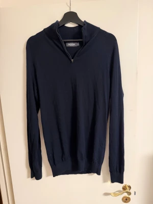 Marinblå merinoulltröja Dressmann M - Mörkblå stickad tröja från Dressmann i mjuk merinoull. Tröjan har lång ärm, ribbade muddar och en hög krage med half zip-dragkedja. Perfekt för dig som gillar stilren och klassisk look.