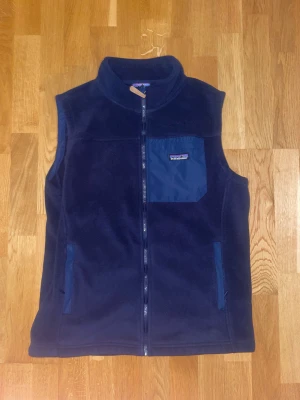 Mörkblå fleeceväst Patagonia - Snygg mörkblå fleeceväst från Patagonia med hög krage och hel dragkedja framtill. Västen har en blå bröstficka med logotyp, samt två sidofickor med dragkedja. Perfekt för lager-på-lager och outdoor. Materialet är mjuk fleece som håller dig varm.