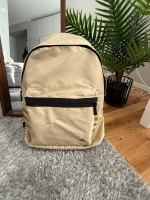 Beige och svart Adidas ryggsäck - Snygg beige ryggsäck från Adidas med svarta detaljer och guldiga ränder på sidan. Framtill finns en stor ficka med svart dragkedja och Adidas-logga i guld. Axelremmarna är vadderade och har både beige och svart färg. 