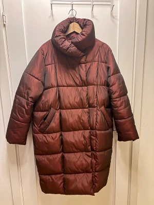 Vinröd pufferkappa från NA-KD strl 38 - Säljer en vinröd, quiltad pufferkappa eller lång jacka från NA-KD i storlek 38. Jackan har hög krage, långa ärmar med ribbade muddar och två sidofickor. Materialet är syntet och jackan har en loose passform som ger en chill vibe. Perfekt för kalla dagar, varm och skön!