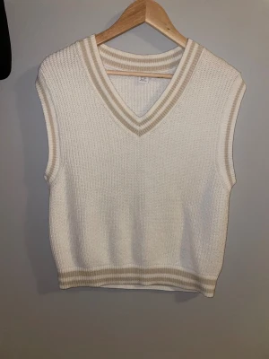 Vit stickad väst med v-ringning - Snygg vit stickad väst med v-ringad hals och ribbade beige ränder vid hals, ärm och nederkant. Den är använd 1-2 gånger. Västen är perfekt att styla över en skjorta eller t-shirt. Enkel och stilren design som funkar året runt. Skriv vid minsta fråga! 