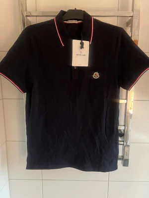 Moncler Pike - Svart pikétröja från Moncler med klassisk krage och korta ärmar. Röd och vit kant på krage och ärmslut ger en snygg kontrast. Broderad Moncler-logga på bröstet. Materialet är bomull och modellen har en normal passform.