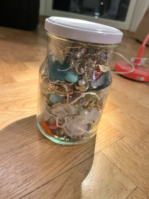 Burk med blandade örhängen och smycken - Stor glasburk “mystiskt” fylld med olika örhängen och smycken i flera färger som silver, guld, blått, rosa, orange och grönt. Innehåller pärlor, metall, plast och tygdetaljer. Perfekt för dig som gillar att mixa och matcha accessoarer och vill ha ett brett utbud av stilar och former. Hela burken väger ca 900 g (se sista bilden) 