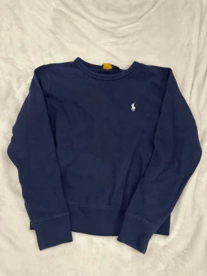 Marinblå sweatshirt från Polo Ralph Lauren - Snygg marinblå sweatshirt från Polo Ralph Lauren med klassisk rund hals och broderad vit logga på bröstet. Tröjan har långa ärmar och ribbade muddar vid ärmslut och nederkant. Perfekt för chill dagar och enkel att matcha med jeans eller joggers.