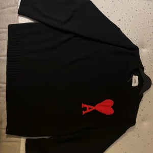 Svart stickad tröja från Ami Paris - Svart stickad tröja från Ami Paris med ikonisk röd hjärtlogga och bokstaven A på bröstet. Tröjan har rund hals, ribbade muddar och lång ärm. Perfekt för dig som gillar stilrena plagg med en twist.