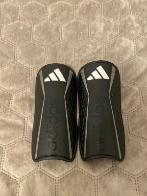 Svarta Adidas benskydd med vit logga - Säljer ett par svarta Adidas benskydd med vit logga och diskreta grå detaljer längs kanterna. Snyggt sportig design med böjd form som sitter skönt på benet. Perfekt för dig som spelar fotboll och vill ha stilrena skydd.