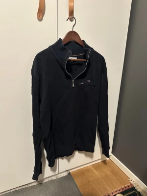 Svart half zip tröja från Bison - Svart långärmad tröja från Bison med ribbad krage och half zip framtill. Tröjan har en liten bröstficka med dragkedja och diskret logga. Materialet är mjuk bomull, perfekt för chill dagar. Snygg och enkel att matcha med jeans eller joggers.