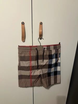 Burberry shorts  - Snygga Burberry shorts med klassiskt rutigt mönster i beige, brun, svart, vit och röd. De har elastisk midja med svart snörning och är tillverkade i lätt polyester. Perfekta för sommaren och har en loose passform som ger en chill vibe. S/XS