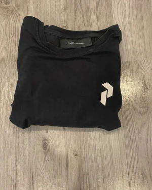 Svart Peak Performance t-shirt i bomull - Snygg svart t-shirt från Peak Performance i storlek L. T-shirten har en klassisk rund hals och ett stilrent vitt logotryck på bröstet. Tillverkad i mjuk bomull som känns skön mot huden. Perfekt för dig som gillar enkel och sportig stil.