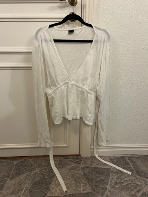 Vit omlottopp med knyt från Gina Tricot - Säljer en vit omlottopp från Gina Tricot i storlek L. Toppen har långa ärmar, djup v-ringning och långa band som knyts i midjan för en snygg siluett. Materialet är lätt och krinklat, vilket ger en chill och luftig känsla. Perfekt för dig som gillar stilrena och enkla plagg.