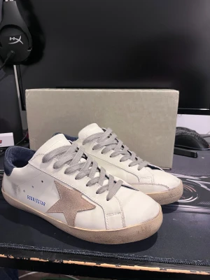 Golden goose  - Säljer ett par GGDB Superstar sneakers i vitt skinn med grå mockastjärna på sidan och grå snörning. Skorna har mörkblå häl och beige sula med svart undersida. Klassisk rund tå och platt sula, perfekt för dig som gillar streetstyle och snygga detaljer. Om priset inte passar ge bud (seriöst)