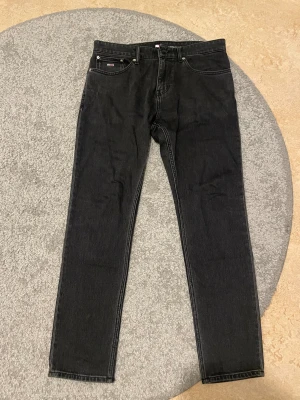 Svarta Tommy Jeans Austin Slim Tapered - Svarta jeans från Tommy Jeans, modell Austin Slim Tapered. Snyggt smal passform med avsmalnande ben, klassisk femficksdesign och ikonisk Tommy Jeans-logga på bakfickan och linningen. Jeansen är tillverkade i slitstarkt denim och har knapp och dragkedja framtill.