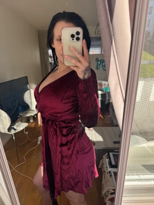 Vinröd omlottklänning (perfekt till jul) NLY Trend - Supersnygg vinröd kort omlottklänning från NLY Trend i glansigt, mjukt material. Klänningen har långa ärmar, v-ringning och knytband i midjan som framhäver figuren. Perfekt för dig som vill sticka ut med en elegant look.