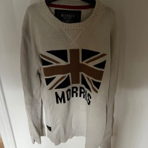Beige stickad tröja Morris M - Snygg beige stickad tröja från Morris i storlek medium. Framsidan har en stor Union Jack-flagga i brunt, svart och vitt samt Morris-tryck under. Tröjan har rund hals och långa ärmar med ribbade muddar. Perfekt för dig som gillar brittisk stil och vill sticka ut.