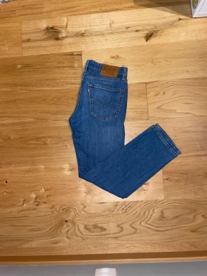 Levi's 511 blå jeans straight fit 30/30 - Säljer ett par klassiska Levi's 511 jeans i blå tvätt med straight fit. Jeansen har fem fickor, knappgylf och brunt läderpatch bak i midjan. Materialet är slitstarkt denim i bomull och passformen är normal med raka ben. Perfekt för dig som gillar tidlös stil.