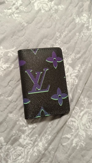 Louis Vuitton svart plånbok med lila mönster - Snygg svart plånbok från Louis Vuitton med ikoniskt LV-märke och lila samt gröna detaljer i klassiskt monogrammönster. Plånboken är i skinn och har flera kortfack på insidan. Perfekt för dig som vill ha något unikt och stilrent i fickan.