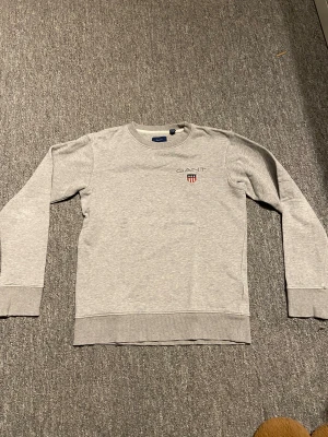 Grå tröja från GANT - Säljer en stilren grå sweatshirt från GANT med broderad logga och sköld på bröstet. Tröjan har rund hals, ribbade muddar och är tillverkad i mjuk bomullsmix. Perfekt för en clean och avslappnad look. Storlek 146-152.