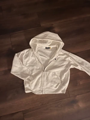 Vit hoodie från Polo Ralph Lauren XL - Vit hoodie från Polo Ralph Lauren i storlek XL. Klassisk design med dragkedja, huva och känguruficka. Broderad svart logga på bröstet. Tillverkad i mjuk bomull, perfekt för chill dagar eller när du vill ha en clean look.
