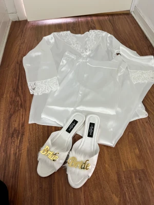 Vita sandaler med Bride-detalj och pyjamas  - Säljer ett par vita sandaler från Ardina Istanbul med öppen tå och platt sula. Framtill har de fluffiga detaljer i syntetpäls och en stor guldig text 'Bride' som sticker ut. Perfekta för brudens morgon eller att chilla i innan festen och pyjamas 