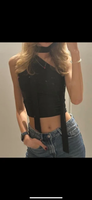 Svart croppad omlott polotopp - Säljer en svart croppad polotopp med omlott-design och oneshoulder. Toppen har långa band som hänger ner framtill och en hög hals som ger en cool vibe. Perfekt att styla med jeans eller kjol för en trendig look.