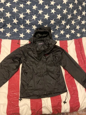 Svart vindjacka från Phelix - Svart vindjacka från Phelix med huva och meshfoder. Jackan har dragkedja framtill, snörning i nederkant och broderad röd logga på bröstet. Materialet är lätt och syntetiskt, perfekt för blåsiga dagar. Snygg och enkel design med lång ärm och praktiska detaljer. Jackan är i gott skick och inte använd så mycket. Jackan är en vind / regnjacka. Barn storlek 150. 