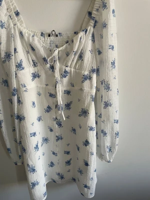 Vit blommig klänning  - Superfin vit kortklänning från Gina Tricot med blått blommönster, långa ärmar med resår och knyt framtill. Klänningen har knappar längs framsidan och ribbad rygg (en liten del). Lätt och luftig med ett extra tyg efter midjan. Klänningen når lite innan knäna. Knapparna går att öppnas. Använt 1 gång. 