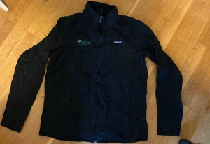 Svart vindjacka från Patagonia - Svart vindjacka från Patagonia med dragkedja framtill och Fairway-brodyr på bröstet. Jackan har ståkrage, lång ärm och en bröstficka med dragkedja. Perfekt för dig som gillar stilren och funktionell design.