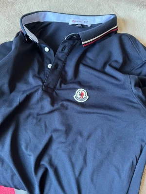 Mörkblå Moncler pikéskjorta - Snygg mörkblå pikéskjorta från Moncler med klassisk krage och vita knappar. Kragen har en stilren detalj med röd och vit rand. Moncler-loggan är broderad på bröstet. Perfekt för dig som gillar exklusiva märken och stilren design.