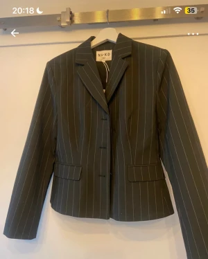 Svart randig kavaj från NA-KD - Snygg svart kavaj från NA-KD med smala vita ränder och klassisk krage. Modellen är figursydd med tre knappar framtill och fickor med lock. Perfekt för dig som gillar stilrena och trendiga plagg med business-vibe.