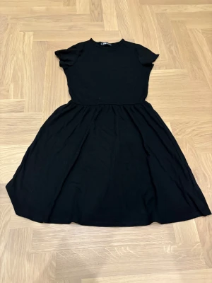 Svart kortklänning från FB Sister - Svart klänning från FB Sister i storlek XS. Modellen har rund hals, korta ärmar och ett utställt kjolparti som ger en skön och avslappnad siluett. Tillverkad i mjuk bomull som känns bekväm mot huden. Perfekt för dig som gillar enkel och stilren design.