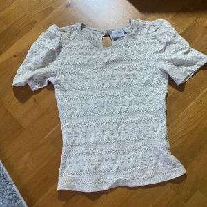 Vit spetsblus från Vila - Säljer en beige blus från Vila med kort puffärm och vacker spets med broderade detaljer. Blusen har rund hals och en liten öppning med knapp i nacken. Perfekt för dig som gillar romantisk stil och fina detaljer.
