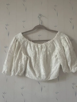 Vit blommig blus med puffärmar - Superfin vit blus med broderade blommor och puffiga korta ärmar. Blusen har en bred, elastisk halsringning. Perfekt för sommarens alla tillfällen!
