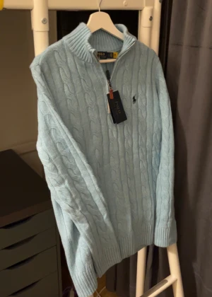 Ljusblå stickad kvartzip från Polo Ralph Lauren kabelstickat - Snygg ljusblå stickad tröja från Polo Ralph Lauren med half zip och klassisk kabelstickning. Tröjan har ribbad krage, muddar och det ikoniska Polo-logot broderat på bröstet. Perfekt för dig som gillar stilren och chill look.
