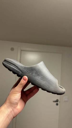Grå Adidas Yeezy Slide sandaler - Säljer ett par grå Adidas Yeezy Slide sandaler med chunky, vågig sula och minimalistisk design. Tillverkade i syntetmaterial som är mjukt och bekvämt för foten. Perfekt för chill dagar och enkel att dra på sig. Diskret Adidas-logga i fotbädden.
