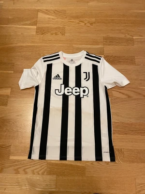 Juventus fotbollströja Adidas svartvit - Juventus matchtröja från Adidas med klassiska svarta och vita ränder, Jeep-logga på bröstet och klubbmärke. Tröjan är kortärmad och tillverkad i lätt funktionsmaterial (AEROREADY) som andas. Perfekt för fotbollsträning eller att bära på läktaren.