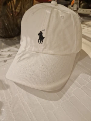 Vit keps från Polo Ralph Lauren - Snygg vit keps från Polo Ralph Lauren med klassisk svart broderad logga framtill och 'POLO' broderat baktill. Justerbar rem med metallspänne för perfekt passform. Tillverkad i mjuk bomull, perfekt för en clean och sportig look.