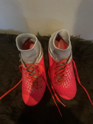 Nike fotbollsskor i rött och grått - Säljer ett par Nike fotbollsskor i en riktigt snygg röd färg med grå detaljer och vita hexagonmönster. Skorna har högt skaft i stretchigt material, snörning och dobbar för gräsplan. Perfekta för dig som vill sticka ut på planen!