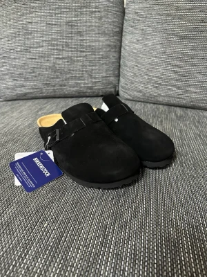 Svarta Birkenstock Boston tofflor - Svarta Birkenstock Boston tofflor i mocka med justerbart spänne på sidan. Skorna har en rund tå, platt sula och innersula i skinn för extra komfort. Yttersulan är i slitstark syntet med mönstrad struktur. Perfekt för chill dagar och enkel stil.