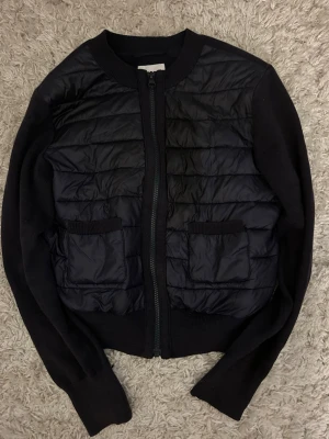 Svart quiltad cardigan jacka Gina Tricot - Svart cardigan jacka från Gina Tricot med quiltad framsida och två fickor. Jackan har dragkedja framtill och är långärmad med ribbade muddar. Perfekt för dig som gillar en stilren look med lite edge.