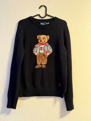 Mörkblå stickad tröja Polo Bear - Mörkblå stickad tröja från Polo Ralph Lauren med ikonisk Polo Bear broderad framtill. Tröjan har rund hals, ribbade muddar och RL-broderi nertill. Perfekt för dig som vill ha en klassisk men lekfull look. Storlek är L men passar böttre rill M