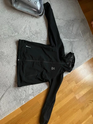 Svart vindjacka från Haglöfs - Svart vindjacka från Haglöfs med huva och hel dragkedja framtill. Jackan är tillverkad i slitstark polyester och har två praktiska fickor med dragkedja. Perfekt för utomhusaktiviteter och har en stilren design med logga på bröstet.