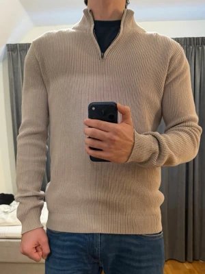 Beige stickad half zip-tröja från John Henric  - Säljer en beige stickad tröja från John Henric med ribbad struktur och half zip-dragkedja i halsen. Tröjan har långärmade och en hög krage som kan bäras öppen eller stängd. Perfekt under värmen! Använd 1 gång, skick 10/10