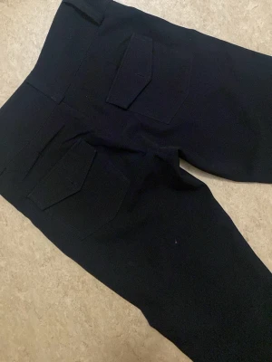 Svarta bootcut-byxor från Gina Tricot - Svarta bootcut-byxor från Gina Tricot i storlek XXS. Byxorna har två knappar fram, fickor med lock bak och är sydda i ett mjukt, stretchigt material som ger en snygg passform. Perfekt för dig som gillar klassisk stil med lite extra edge. Aldrig använt för att är för små.
