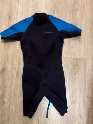 Olaian våtdräkt med blå detaljer - Kortärmad våtdräkt från Olaian i mörkblått med ljusblå axelpartier. Dräkten har dragkedja bak med lång rem för enkel stängning. Perfekt för vattenaktiviteter och har mått markerade på bilderna för passform. Materialet är syntetiskt och designen är sportig.