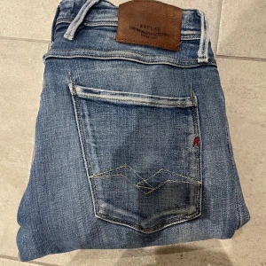 Replay blå jeans - Säljer ett par snygga blå Replay jeans i modellen anbass och  i storlek 33/32. I  De har klassisk femficksdesign, slitningar och ett hål på ena knät för en cool look. Jeansen har raka ben och tydliga kontrastsömmar. Märkespatch i läder baktill i midjan.